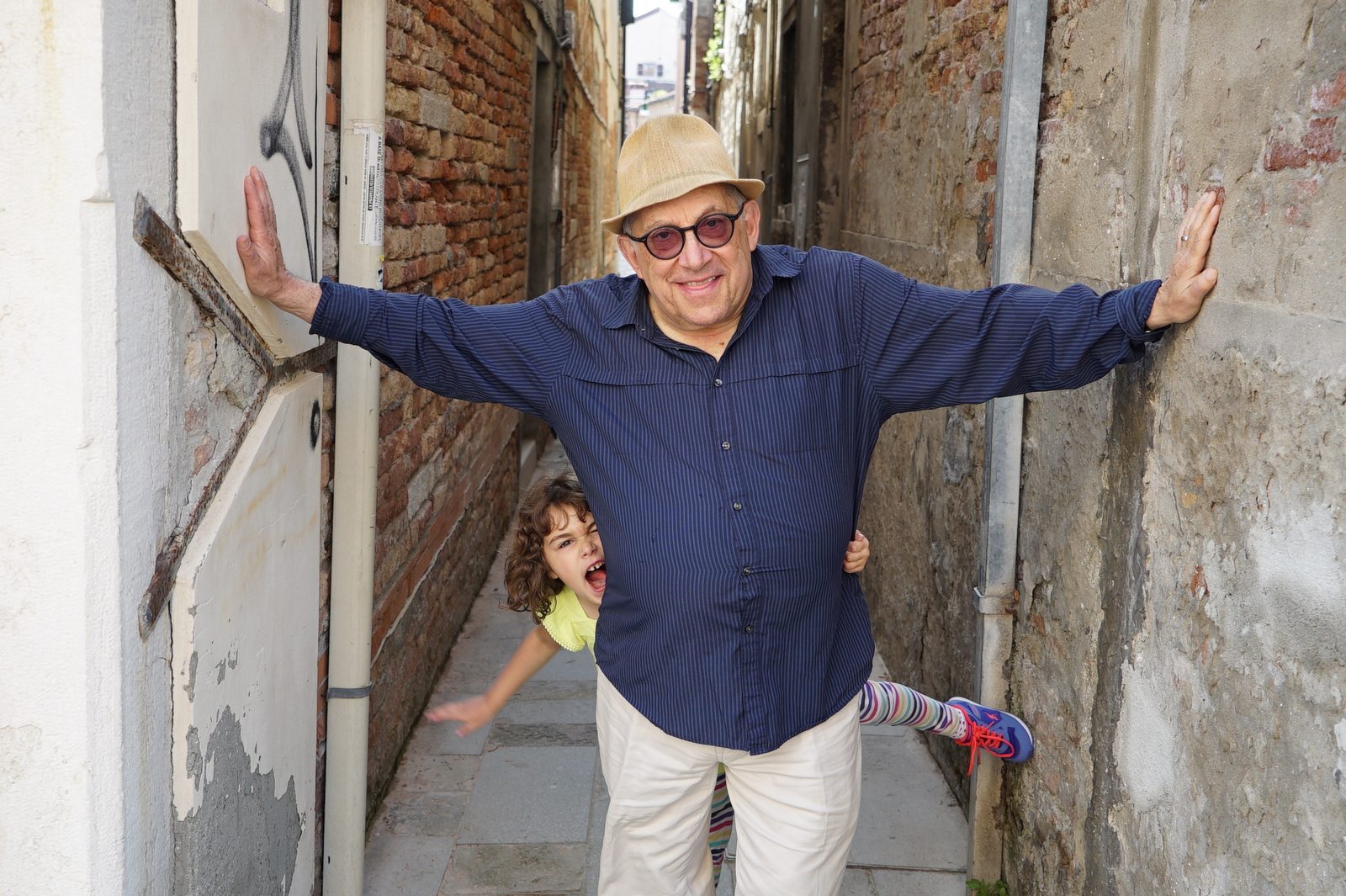 David stretching his arms across a narrow brick alley in Venice while a child ducks out from behind him