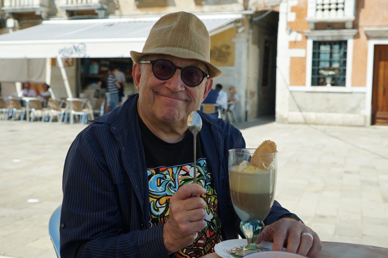 David in a straw hat and tinted glasses smiling over a tall coffee dessert and a long spoon
