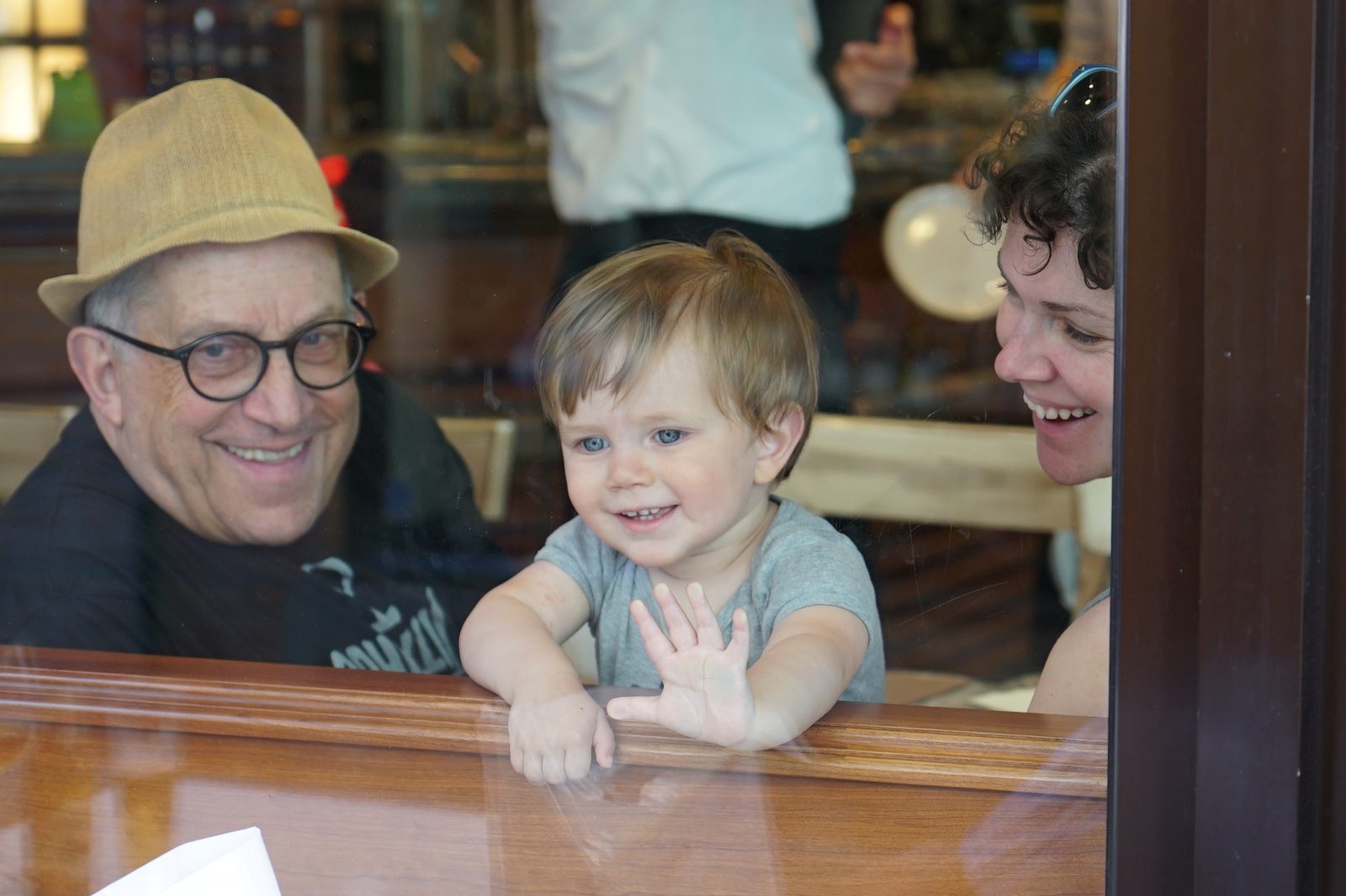 David smiling beside a waving toddler and a woman, seen through a restaurant window