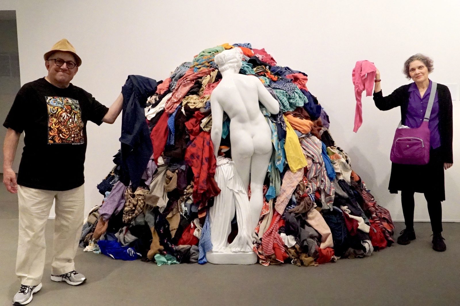 David and a woman holding up clothes beside a huge pile of fabric with a white statue at a museum