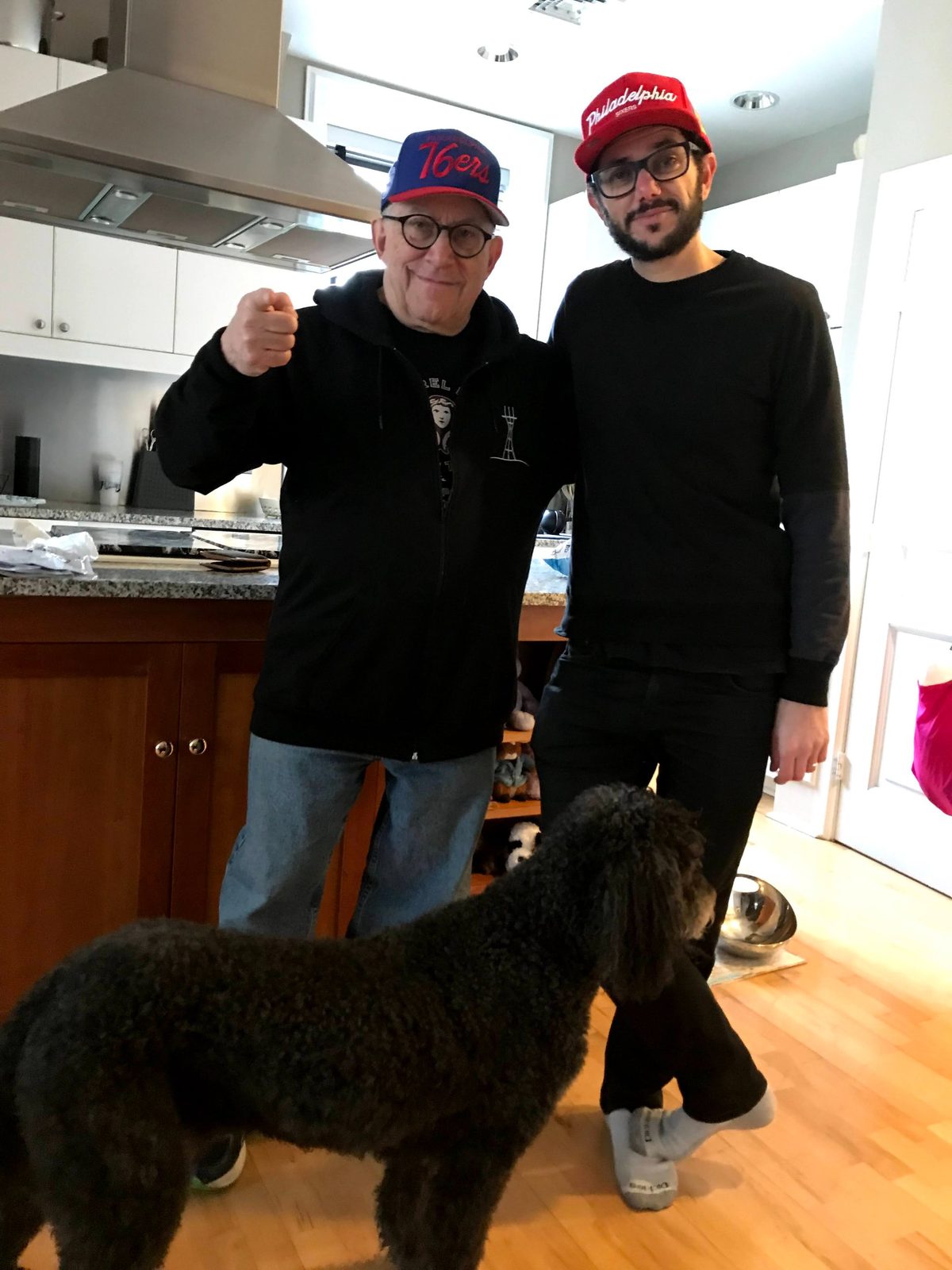 David and a younger man in 76ers and Philadelphia caps with a black poodle in a kitchen