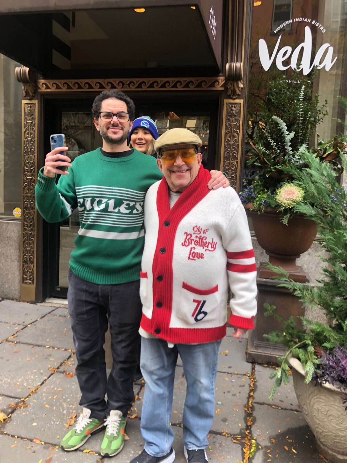 David in a red and white 76ers cardigan beside a man in an Eagles sweater outside a restaurant