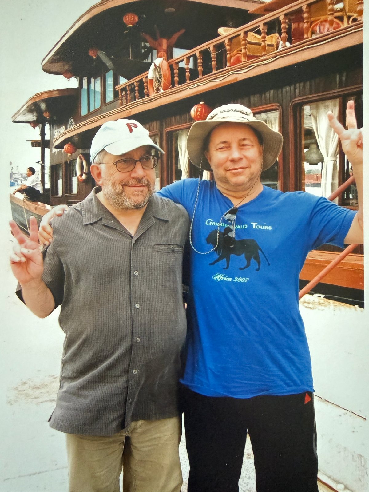 David in a Phillies cap beside a friend in a Greenwald Tours Africa 2007 shirt, both making peace signs in front of a river boat