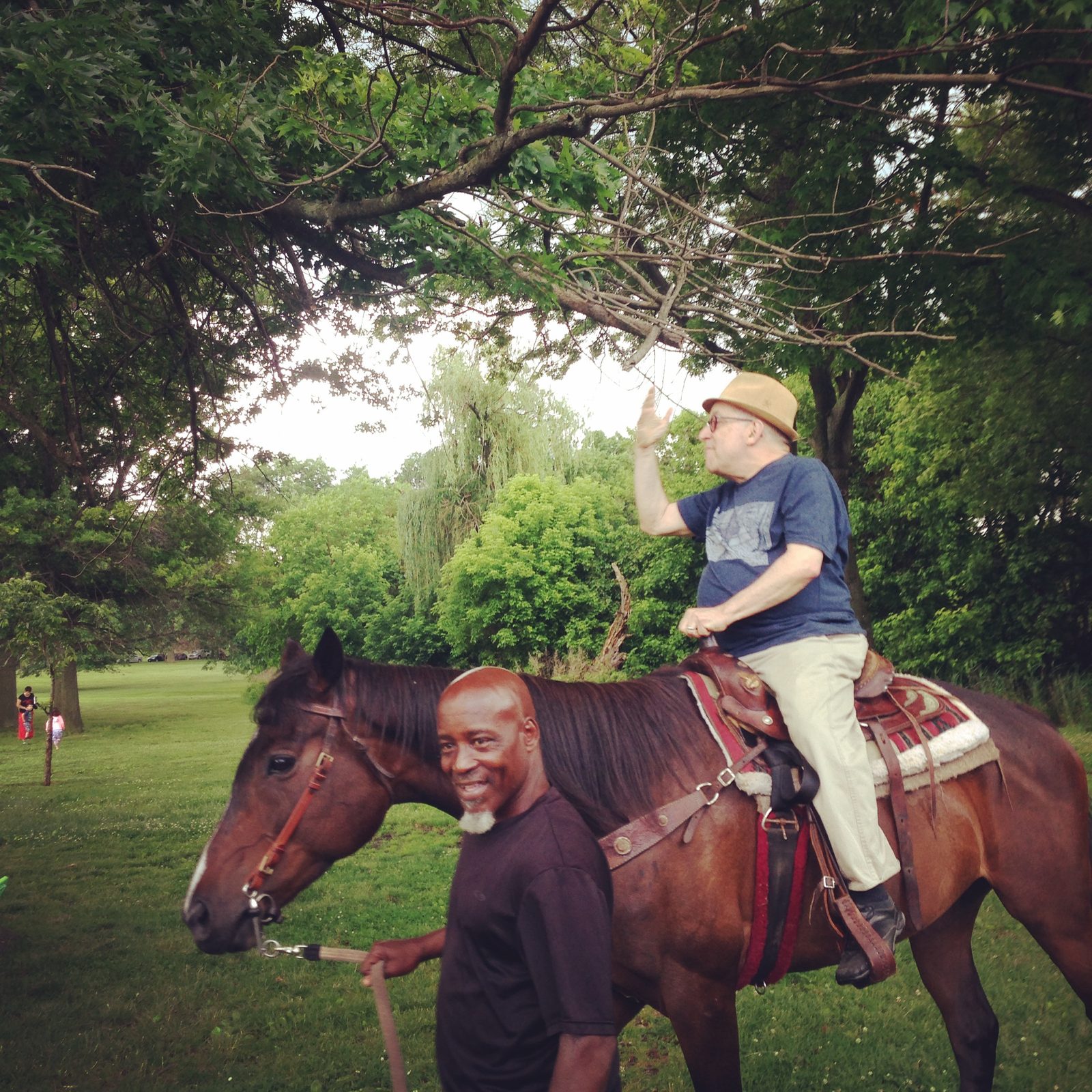 David in a straw hat waving from a horse in a park while a man leads it