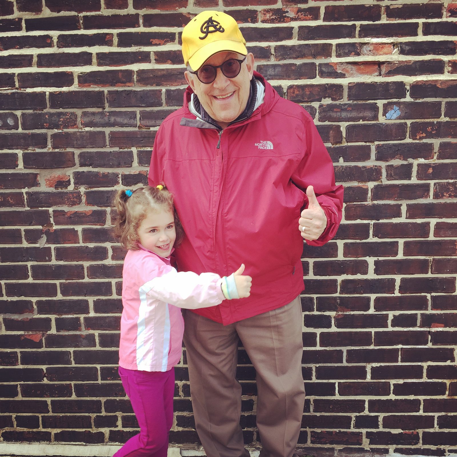 David in a yellow cap and red jacket giving a thumbs up with a young girl hugging him in front of a brick wall