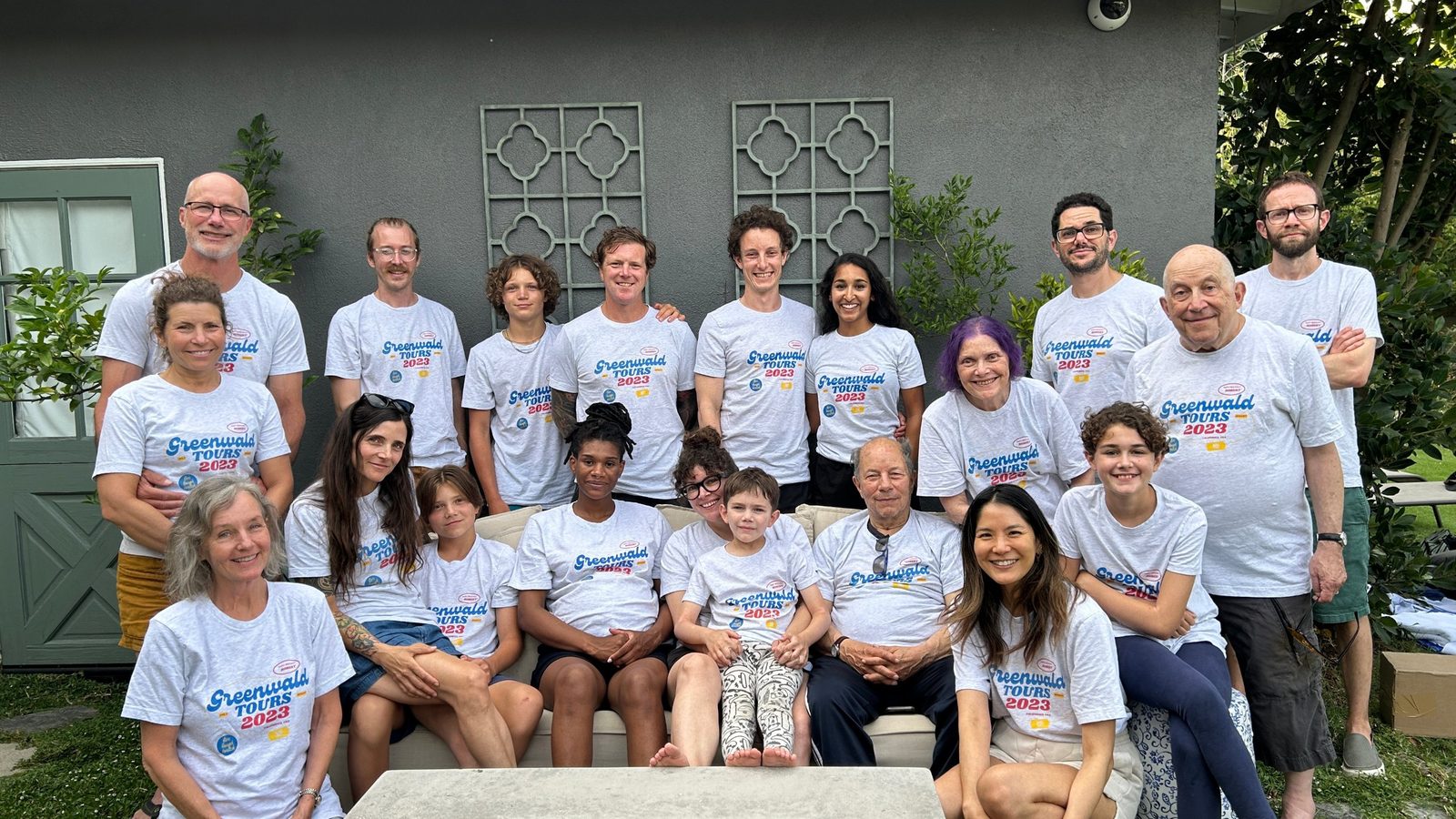 A large family group in matching Greenwald Tours 2023 shirts outside a gray house