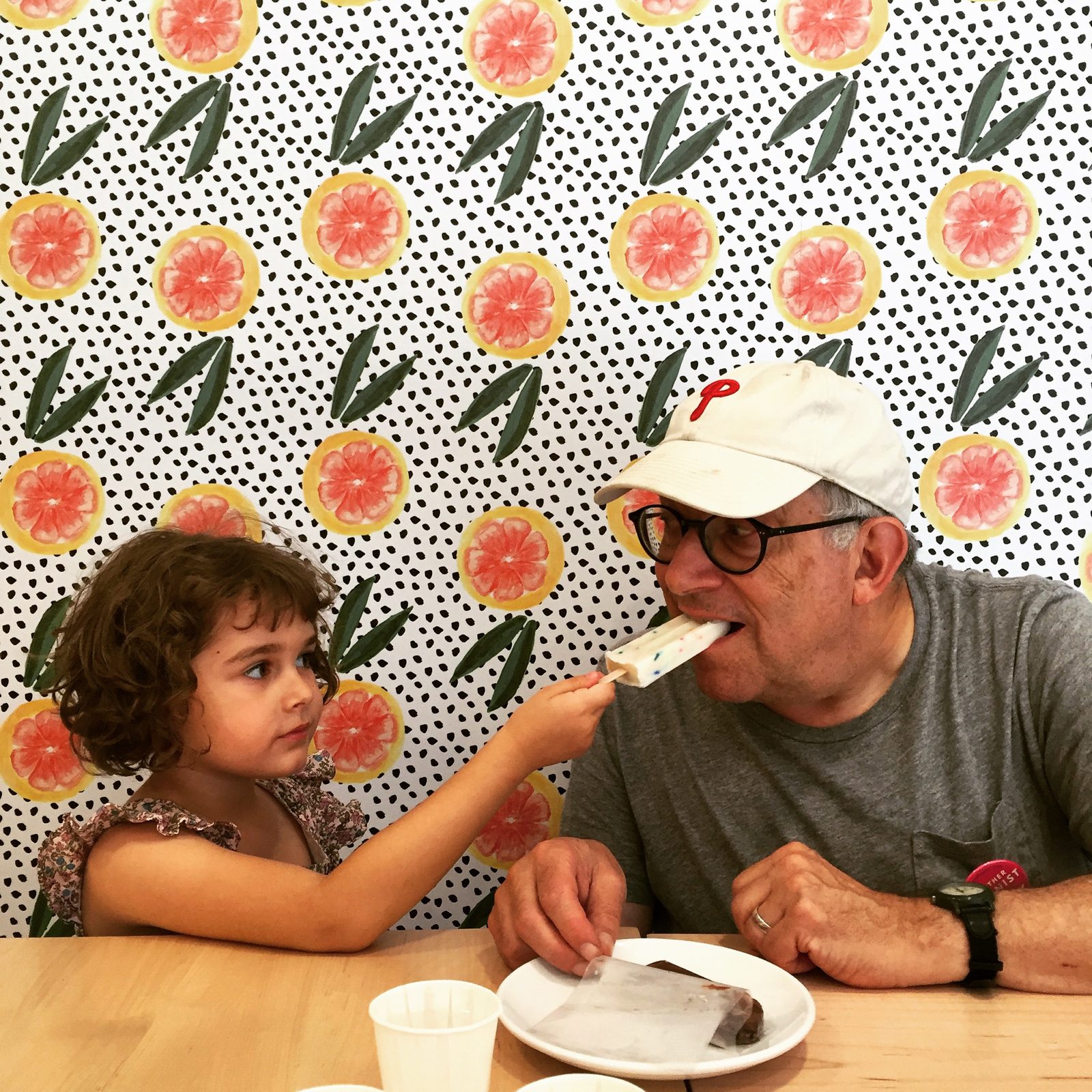 A young girl feeding David a popsicle in front of a grapefruit-pattern wall