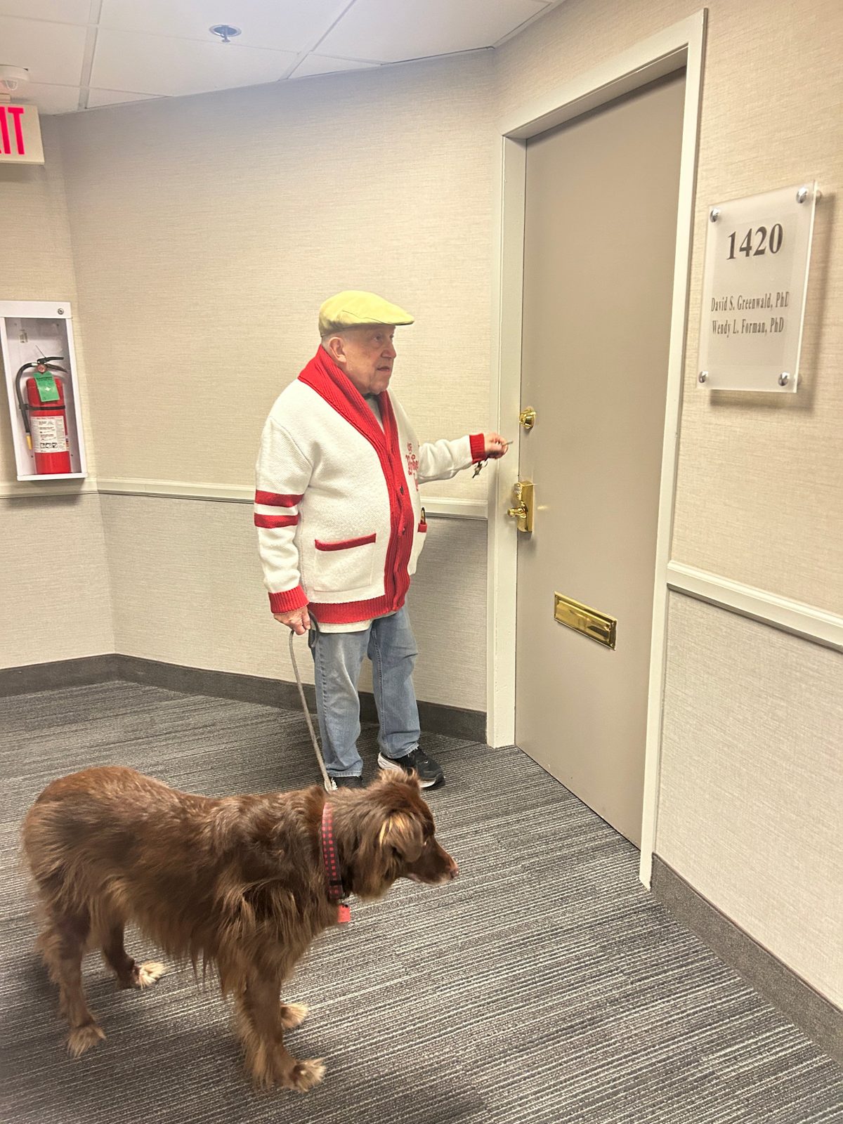 David in a red and white cardigan unlocking an office door with a brown dog beside him