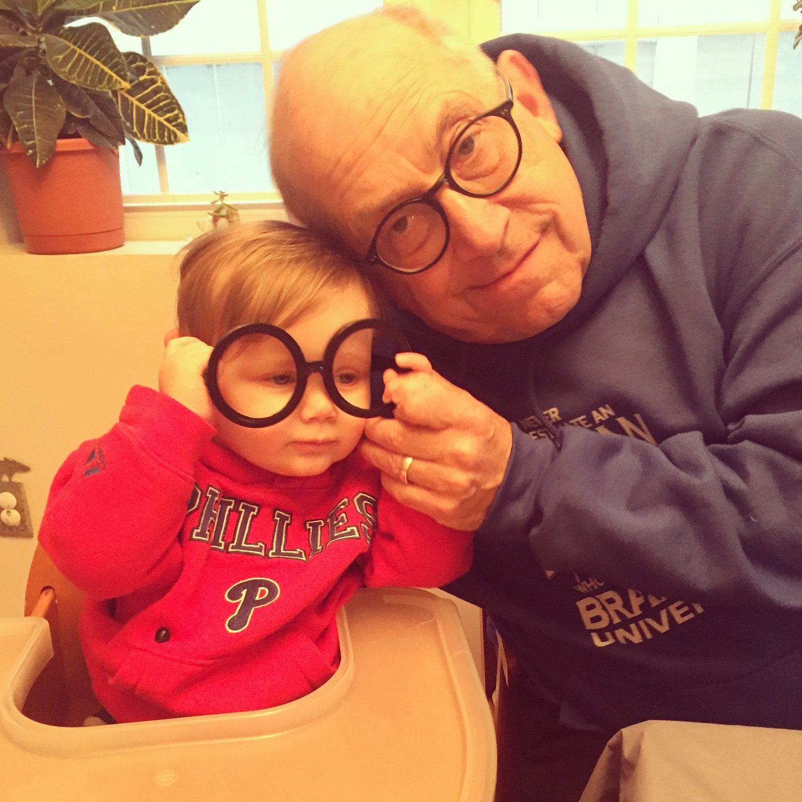 David leaning his head against a toddler who holds up a pair of big round black glasses, the toddler in a Phillies sweatshirt