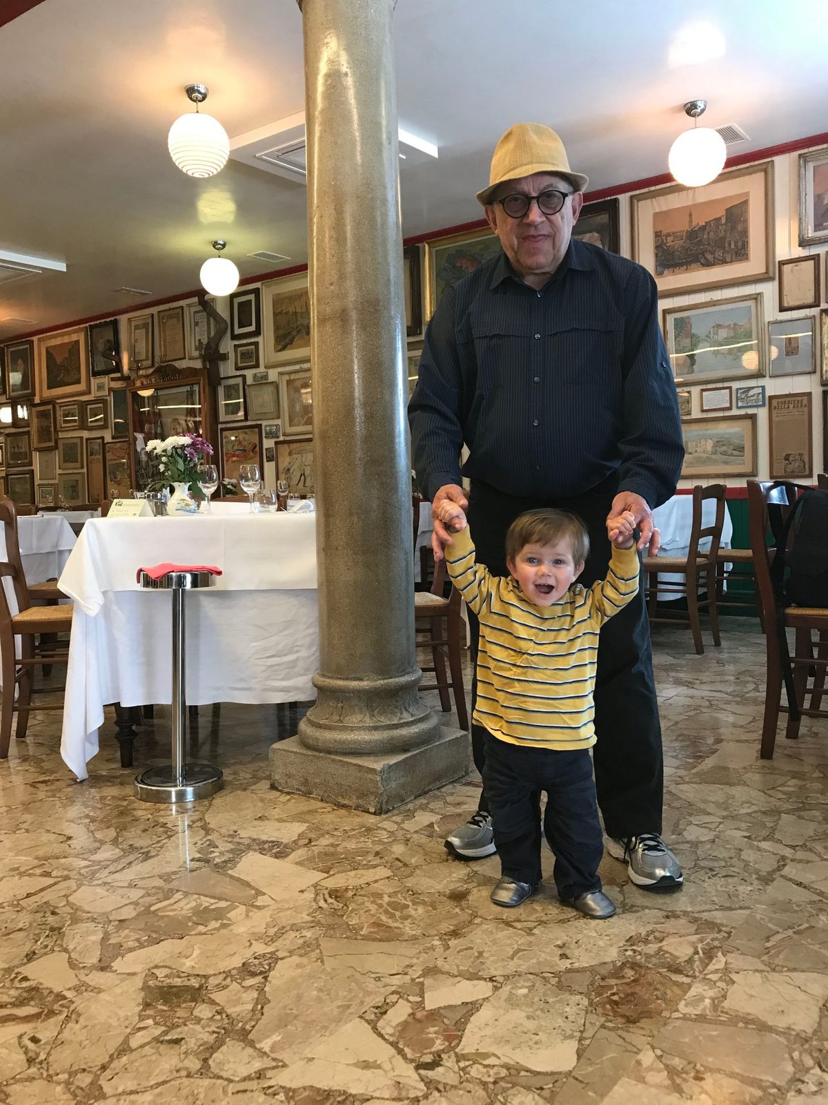 David in a straw hat holding the hands of a toddler in a yellow striped shirt in an Italian restaurant