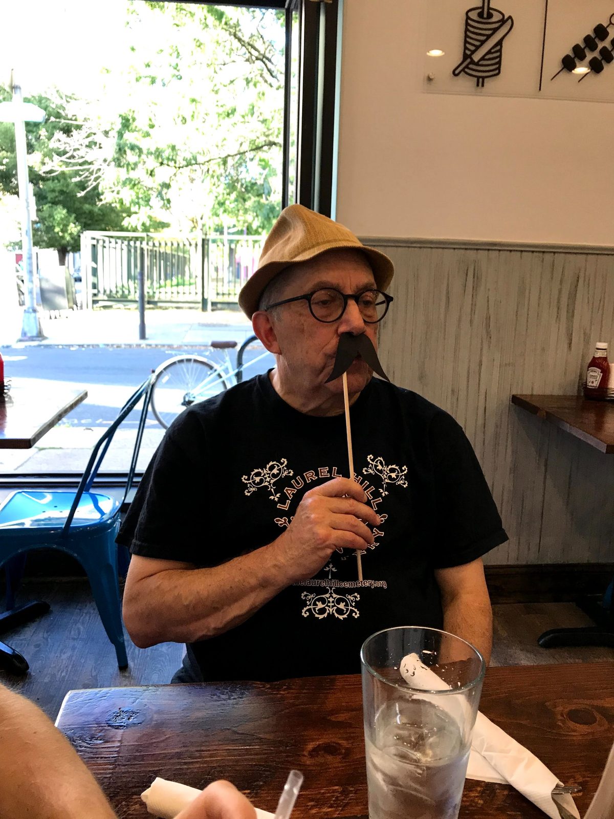 David in a straw hat holding a paper mustache on a stick under his nose at a restaurant table