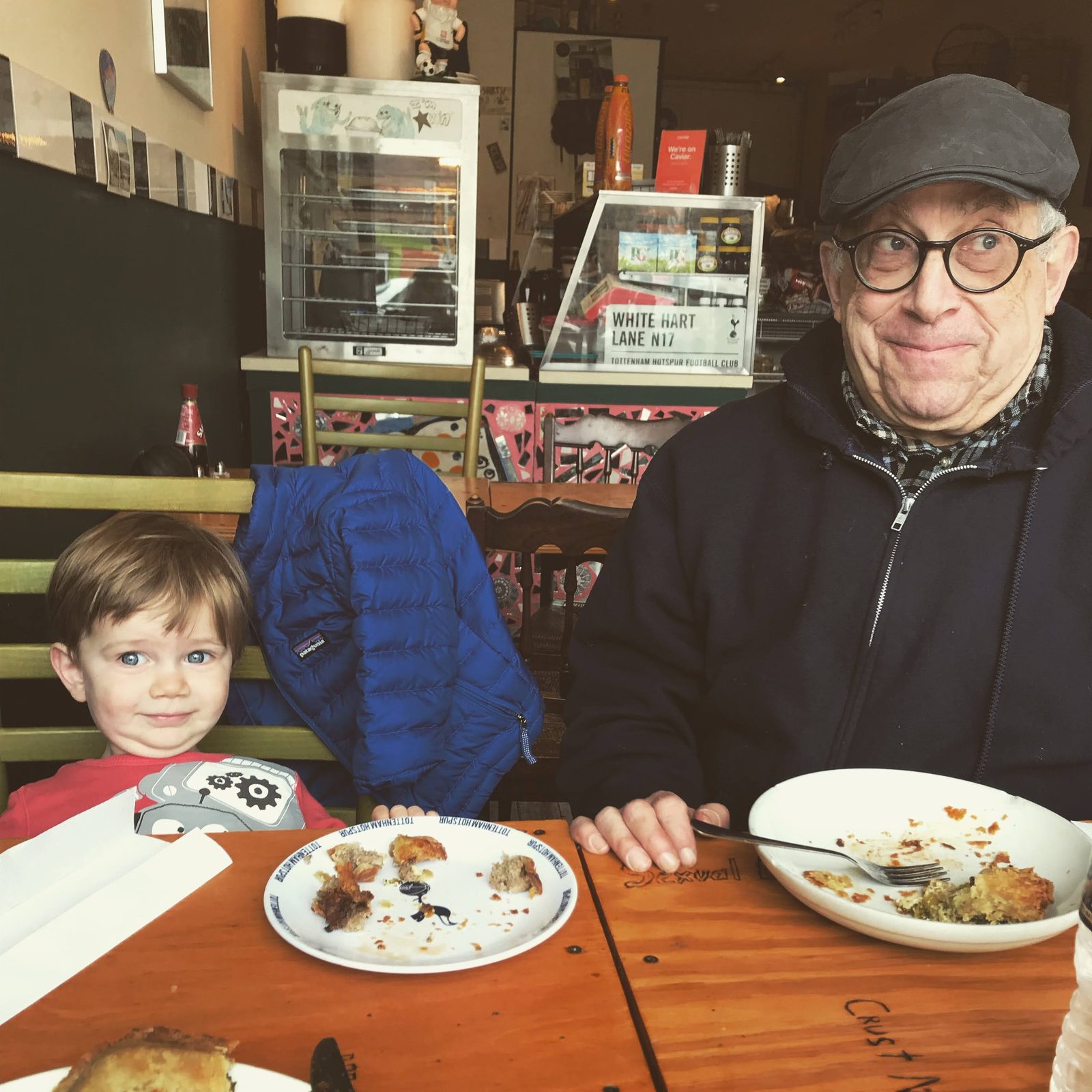 David in a flat cap making a wry face next to a toddler at a cafe table