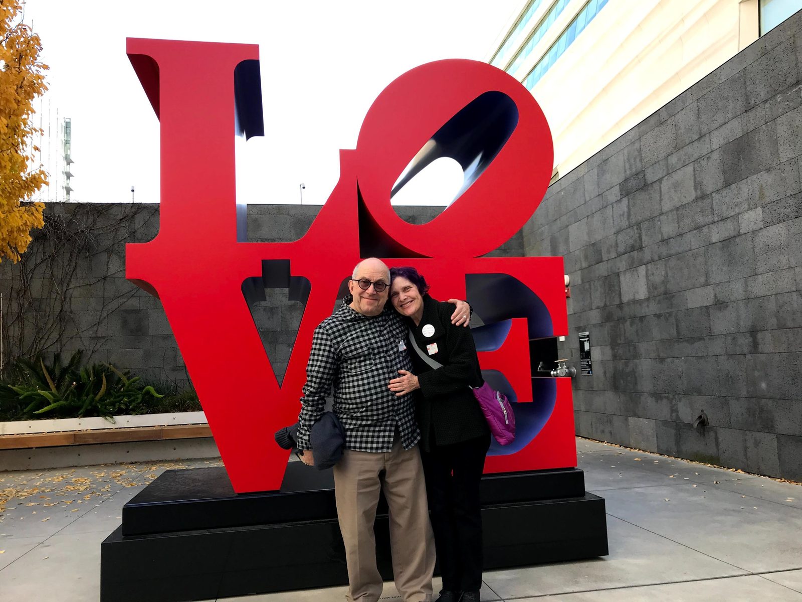 David and a woman with purple hair embracing in front of the red LOVE sculpture
