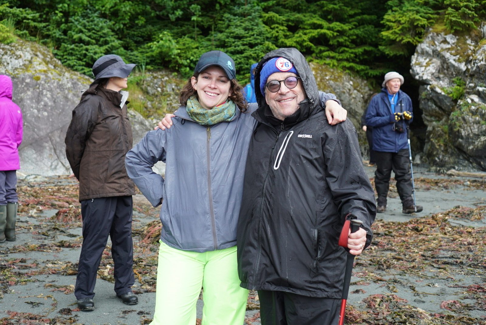 David in a 76ers beanie and rain jacket with his arm around a woman on a rocky Alaska beach