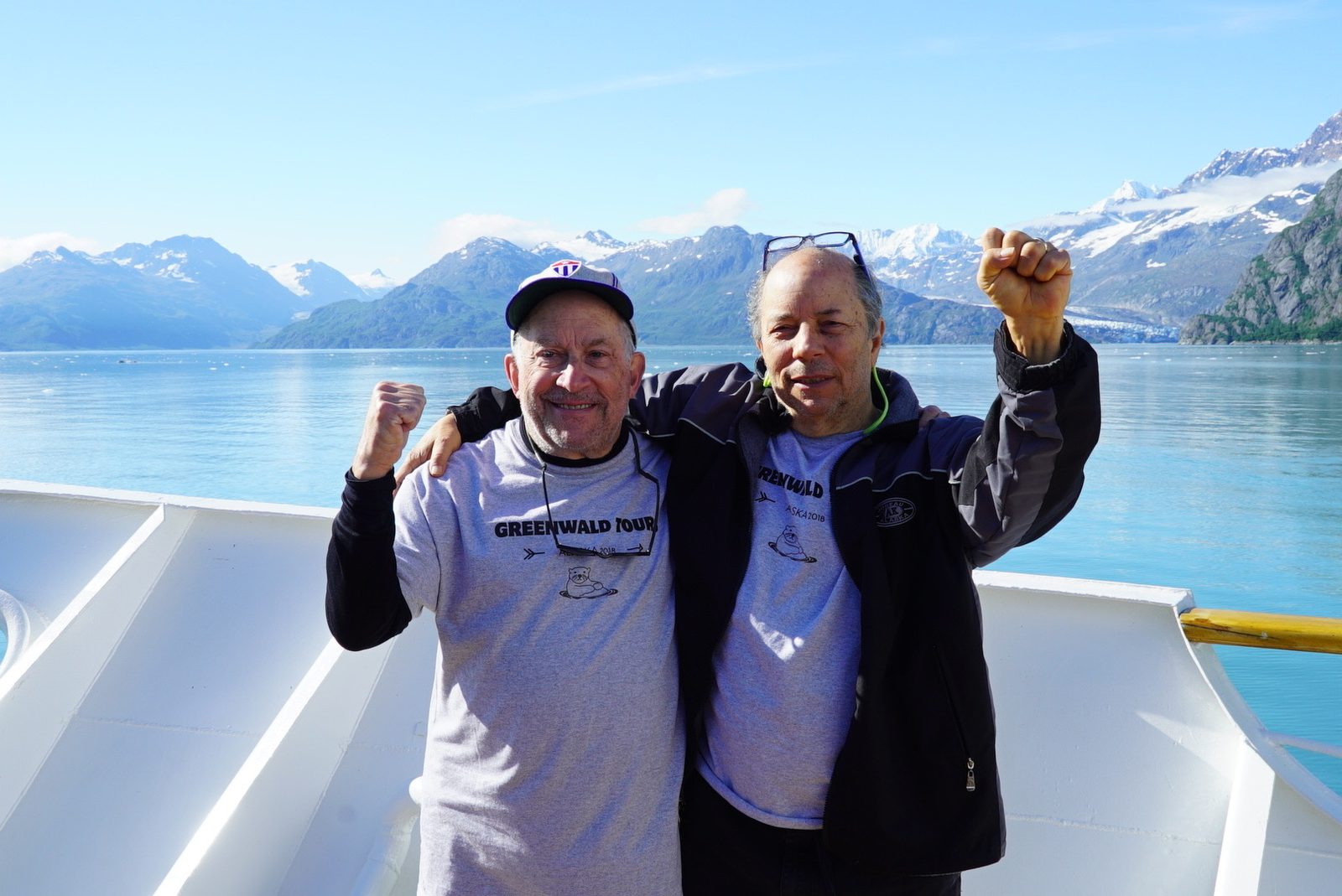 David and a man raising their fists in matching Greenwald Tours Alaska 2018 shirts on a boat in front of mountains
